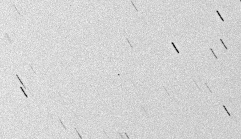 The image shows the NASA Mars 2020 spacecraft against a background of stars. The image is a negative view with the sky white and the stars and spacecraft dark. The spacecraft is a small dark spot in the center of the image. The telescope tracked the spacecraft which is why the stars are streaked from upper left to lower right.